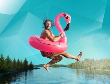 Mann in Badeklamotten springt in einem aufblasbaren Flamingo in den See